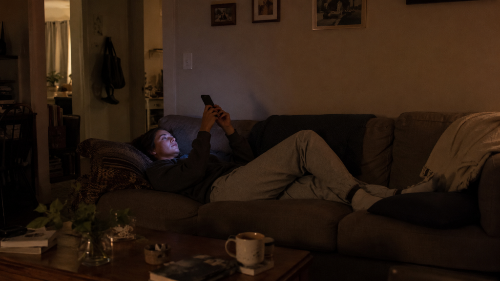 Woman lying on a couch in a dark living room, face lit by phone screen, scrolling in the evening.